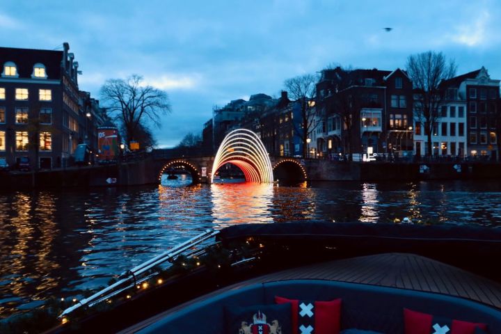 a bridge with light Amsterdam Light Festival artwork over a body of water with a Amsterdam city in the background